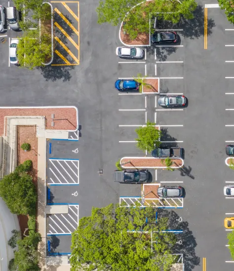 Aerial view of a parking lot with marked spaces and handicap spots, several cars parked, with trees and a building nearby.